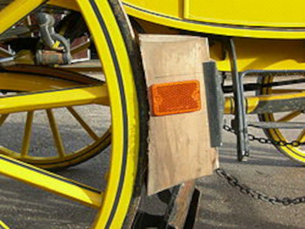 Жёлтое деревянное колесо конного экипажа, к которому прижата деревянная колодка-башмак. К колодке прикреплён современный оранжевый светоотражатель.