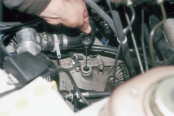 На изображении часть моторного отсека автомобиля, за шлангами виден корпус механической коробки передач. Мужская рука держит щуп, наполовину вынутый из маслозаливного отверстия.